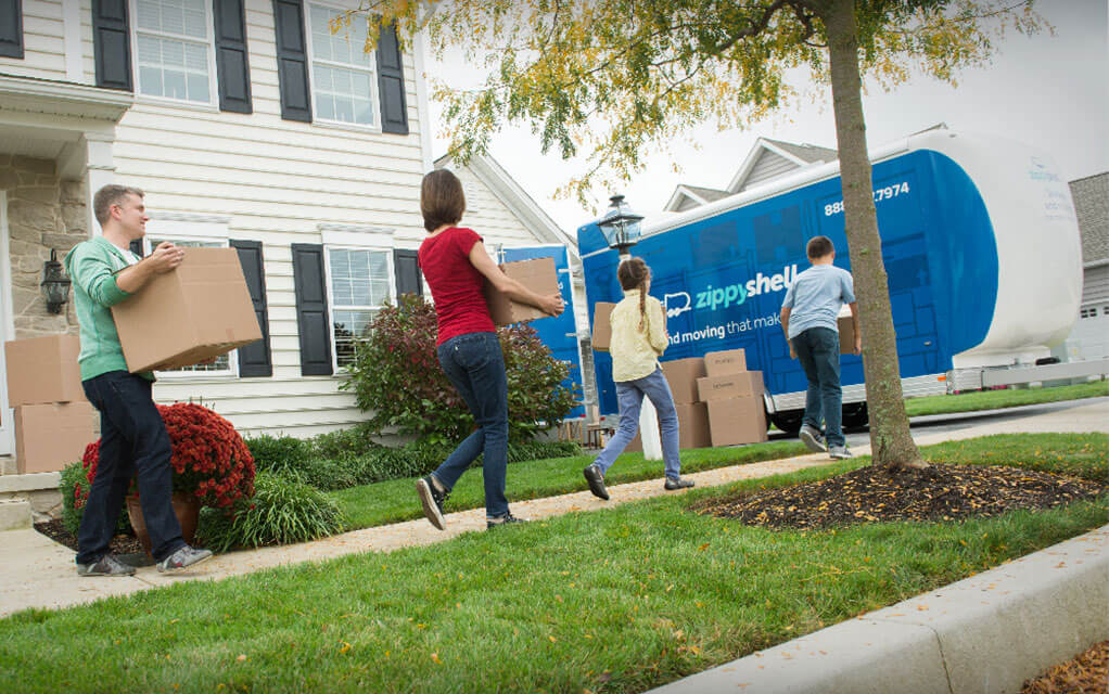 A suburban family carries boxes out to the Zippy Shell container that is parked in their driveway as they get their move started
