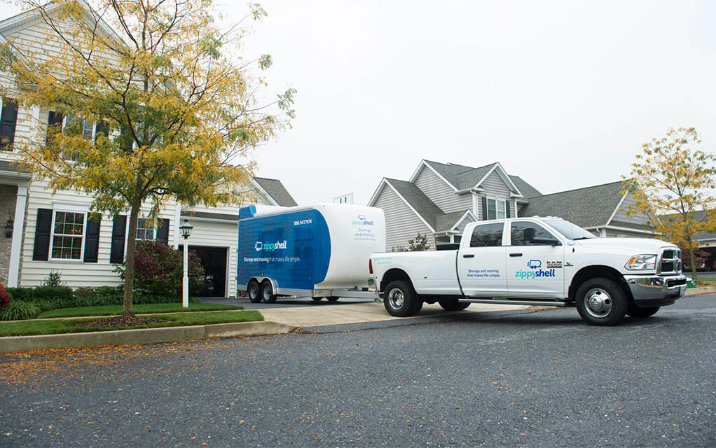 A Zippy Shell truck backs a portable storage container into the driveway of a suburban home on a cloudy day