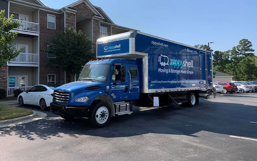 A blue Zippy Shell box truck sits in the parking lot of an apartment complex waiting to be packed up for a move